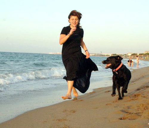 Η Ιωάννα Γκέρτσου σε νέα ηλικία, φορά ένα μαύρο φόρεμα και τρέχει δίπλα στη θάλασσα με το μαύρο λαμπραντόρ της
