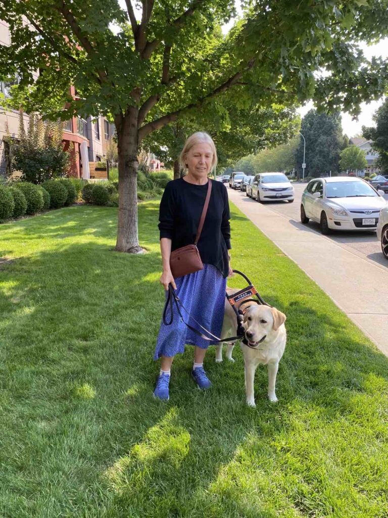 Η Λιάνα, μια ξανθιά κυρία με μπλε φούστα και μπλε αθλητικά παπούτσια και μαύρο φούτερ, με τον σκύλο οδηγό της, ένα ξανθό λαμπραντόρ.